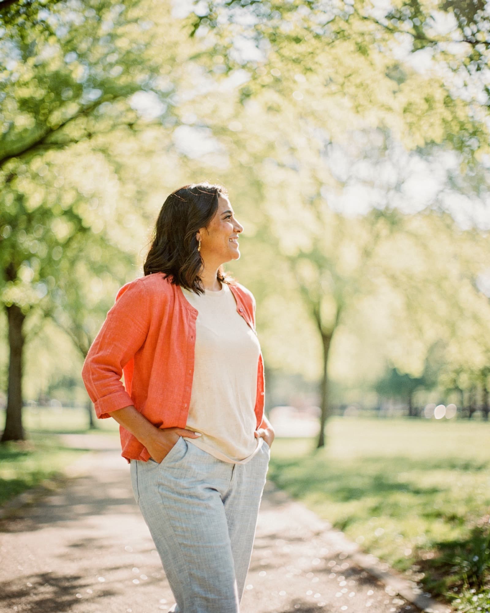 Woman standing on a sunlit park path in late spring, hands in her pockets, looking off toward the trees