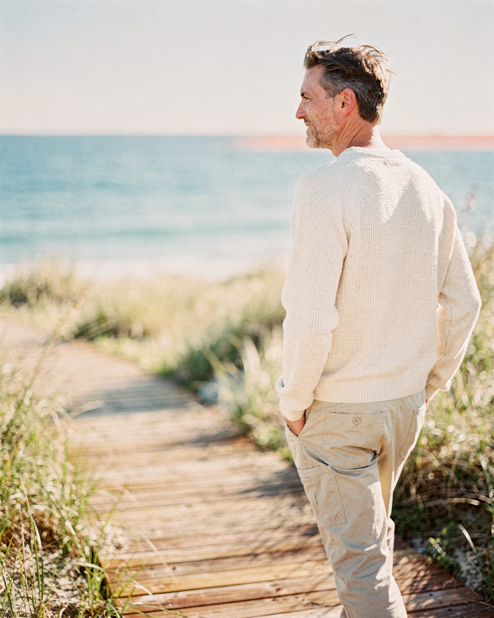 Man walking on a coastal boardwalk along sunlit dune grass at morning