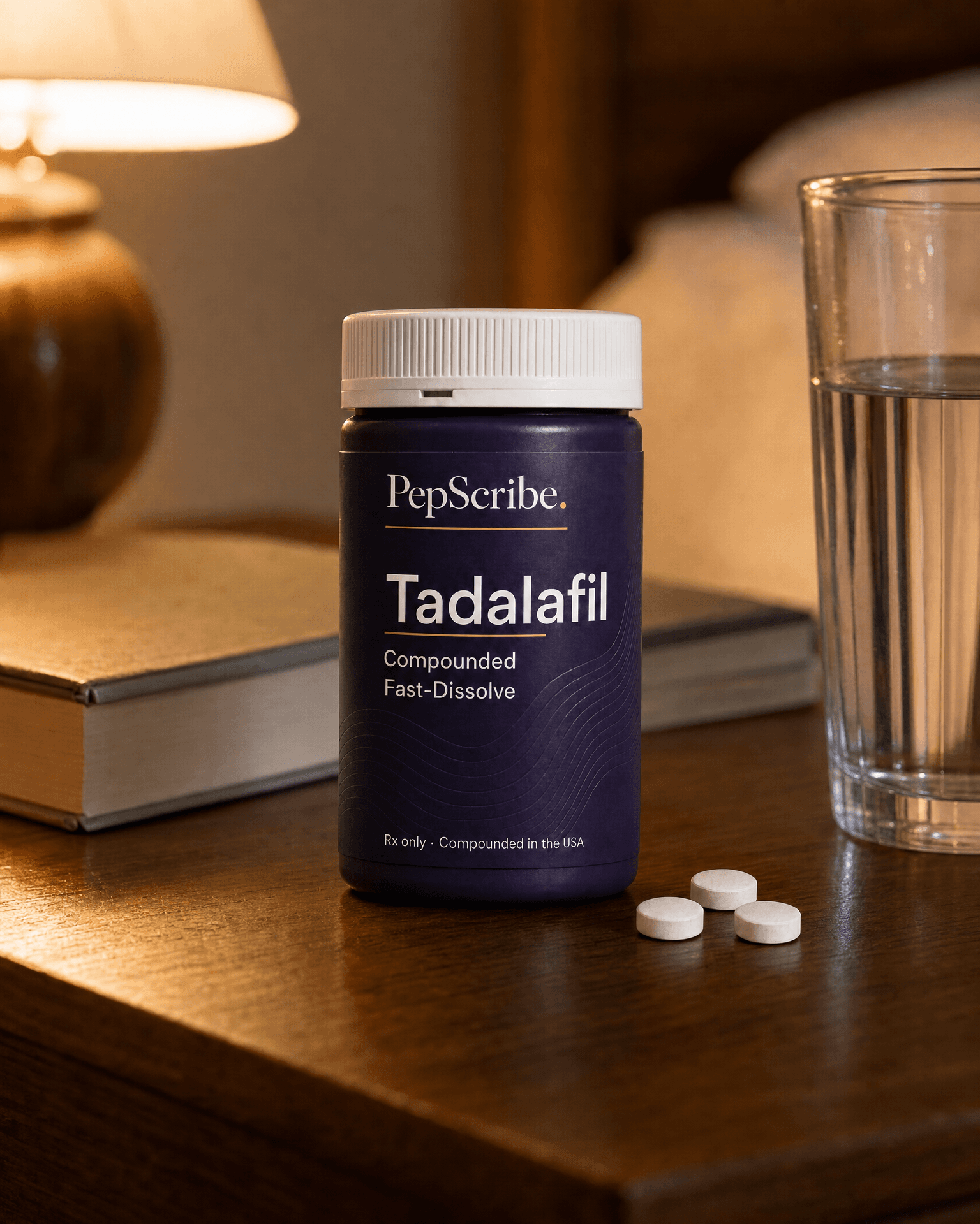 A PepScribe-branded compounded fast-dissolve tadalafil tablet on a warm walnut nightstand beside a lamp, a book and a glass of water.