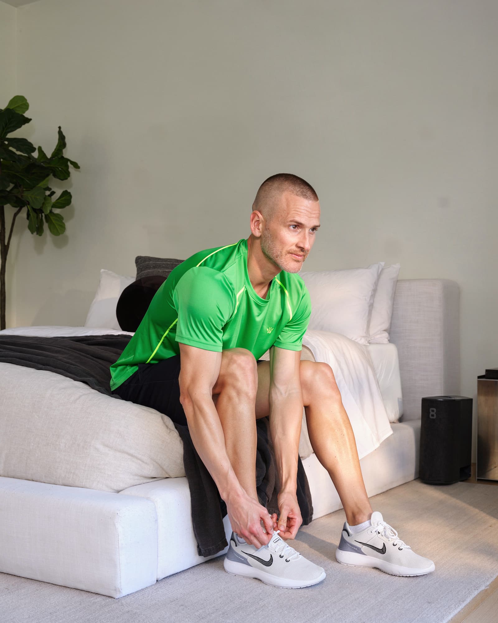 A fit American man in his late thirties sitting on the edge of the bed lacing a shoe as he wakes, energetic and healthy.