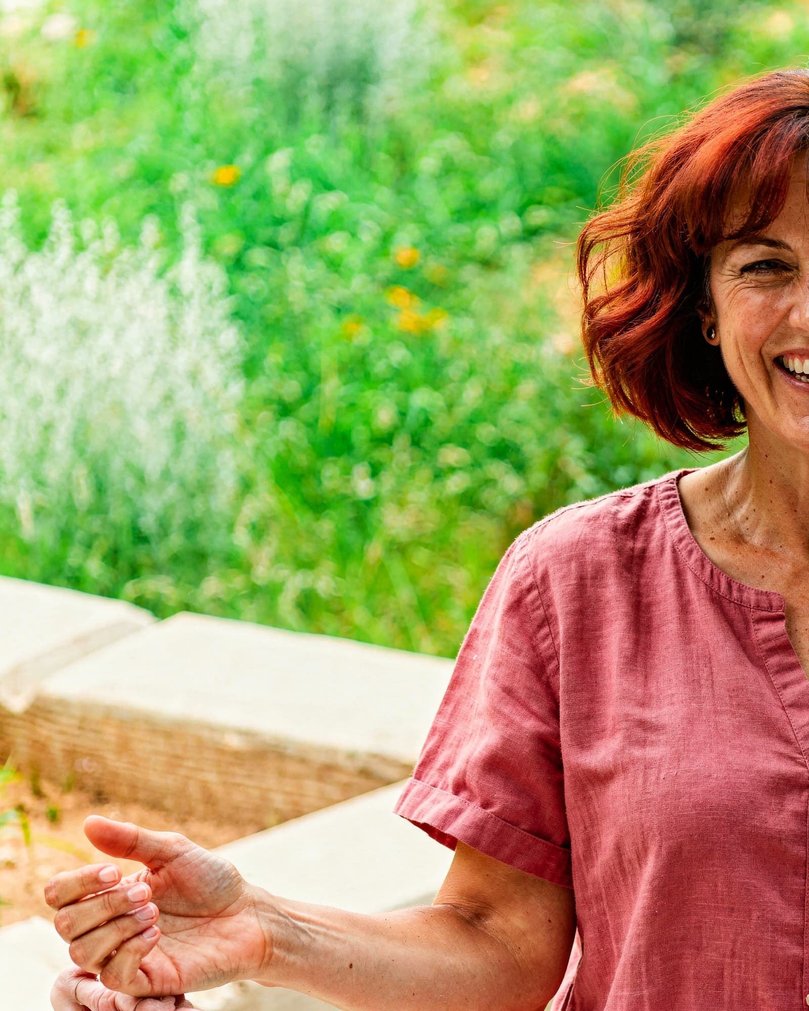An American woman in her late forties laughing in her lush garden, warm and energetic.