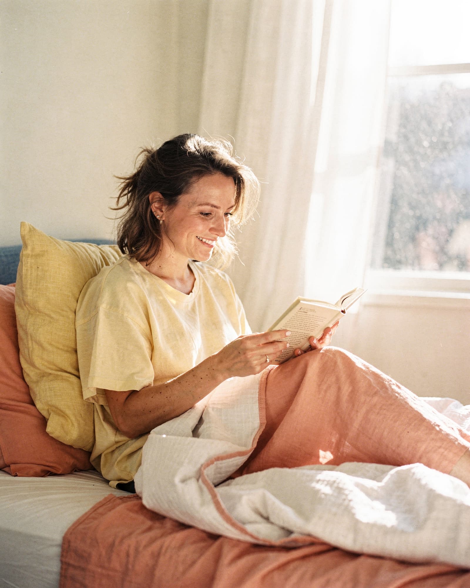 Woman reading a paperback book in a sun-drenched bedroom